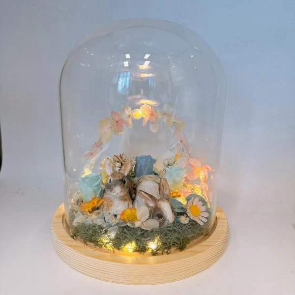 一座玻璃罩擺飾置於淺色木質圓形底座上，罩內鋪有苔蘚與碎石，中央有兩隻兔子動物公仔相依而坐；周圍點綴多朵永生花與小雛菊、藍色與粉色玫瑰，後方以粉橘色系花瓣組成半圓拱門。底座四周有暖黃小燈串發光，整體呈溫暖柔和的夜燈效果。