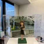 背後襯托著校舍照片，整體效果極具紀念意義。 公仔造型簡約典雅，身穿 黑色過膝連身裙，腳踏深色平底鞋，手中抱著書本，營造出 知性與專業 的氣質。細緻雕塑的臉龐帶著微笑，搭配標誌性的 及肩直髮與眼鏡造型，塑造出親切、可靠的形象。 底座以 綠色草地質感 呈現，呼應校園氛圍；透明保護盒讓公仔能被長久完整保存，防塵又具展示價值。這款設計特別適合作為 教師禮物、退休紀念、畢業送別或校園回憶收藏，帶有濃厚的紀念感與專屬心意。