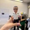 客製化個人職人公仔——制服警務人員/保安人員 手工人物公仔與細節 （圖片展示了一尊被手持著的客製化Q版軟陶公仔，主角是一位穿著制服的中年男性。公仔同樣採用頭大身小的Q版風格，面部特徵刻畫細膩，有著灰白的短髮和平頭造型，戴著黑框眼鏡，表情沈穩嚴肅，嘴角微微抿起。他身穿標準的白色短袖制服襯衫，肩上有肩章，胸前有口袋及名牌（依稀可見名字為「CHOW MING TAK」），下身穿著黑色長褲與黑皮鞋。他的姿勢是右手插腰，左手夾著一頂黑色大盤帽（帽沿有紅色線條裝飾），展現出執勤時的標準儀態。公仔固定在一個簡單的灰色圓形底座上。） 環境背景與光影 （這張照片是在室內拍攝的，背景是一間典型的辦公室或工作間。天花板上有明亮的長條形日光燈管，提供了均勻且充足的冷白光照明。背景處於失焦狀態（景深模糊），可以看到白板上貼有一些紙張或圖表，還有辦公桌、電腦螢幕以及雜物堆疊的角落，顯示出這是一個真實的工作環境。一隻手從左下方伸入鏡頭，穩穩地握住公仔的腿部及底座，將其展示在畫面中央。） 客製化公仔、Q版人像、制服公仔、警察/保安、退休禮物、榮休紀念、CHOW MING TAK、大盤帽、手工軟陶、專屬訂製 嚴肅專業、盡忠職守、正直穩重、職業榮譽、紀念價值、辦公室擺設、長官風範、制服細節