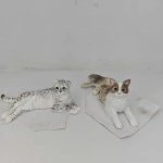 兩隻小型貓仔公仔置於淺色背景與紙片上：左側為銀灰虎斑花紋、半躺側身、尾巴向後蜷起，臉部圓潤眼神明亮；右側為白色搭配棕色斑塊、前腳向前趴姿、尾巴自然伸展。兩款皆以細緻刻劃的毛流與陰影塗裝呈現，體現成對展示風格。