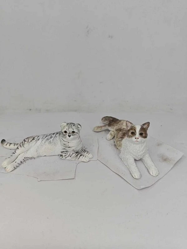 兩隻小型貓仔公仔置於淺色背景與紙片上：左側為銀灰虎斑花紋、半躺側身、尾巴向後蜷起，臉部圓潤眼神明亮；右側為白色搭配棕色斑塊、前腳向前趴姿、尾巴自然伸展。兩款皆以細緻刻劃的毛流與陰影塗裝呈現，體現成對展示風格。