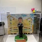 一座透明展示盒中的學校人像公仔：角色穿黑色西裝與條紋領帶，雙手抱臂站在綠色草地質感的圓角底座上。前方橘色名牌寫有英文字樣，向受贈者致謝並祝福退休。腳邊放有高爾夫球與球座，以及一個裝著綠色物件的小籃子。背板為米黃色多窗校園建築，天空藍白雲為背景，整體置於室內桌面拍攝。