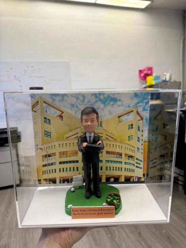 一座透明展示盒中的學校人像公仔：角色穿黑色西裝與條紋領帶，雙手抱臂站在綠色草地質感的圓角底座上。前方橘色名牌寫有英文字樣，向受贈者致謝並祝福退休。腳邊放有高爾夫球與球座，以及一個裝著綠色物件的小籃子。背板為米黃色多窗校園建築，天空藍白雲為背景，整體置於室內桌面拍攝。