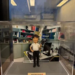 透明壓克力展示盒內的商業人像公仔：角色身穿白色短袖襯衫與黑色長褲，單手插腰、另一手比讚站立於灰色底座前。背板是堆滿文件夾、書籍與電腦螢幕的辦公室工作區。前方金色銘牌寫有「Happy Boss, Kin phar ♥ Love from RTSKH Pharmacy」訊息。