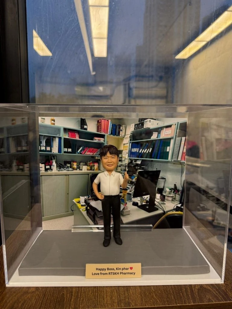 透明壓克力展示盒內的商業人像公仔：角色身穿白色短袖襯衫與黑色長褲，單手插腰、另一手比讚站立於灰色底座前。背板是堆滿文件夾、書籍與電腦螢幕的辦公室工作區。前方金色銘牌寫有「Happy Boss, Kin phar ♥ Love from RTSKH Pharmacy」訊息。
