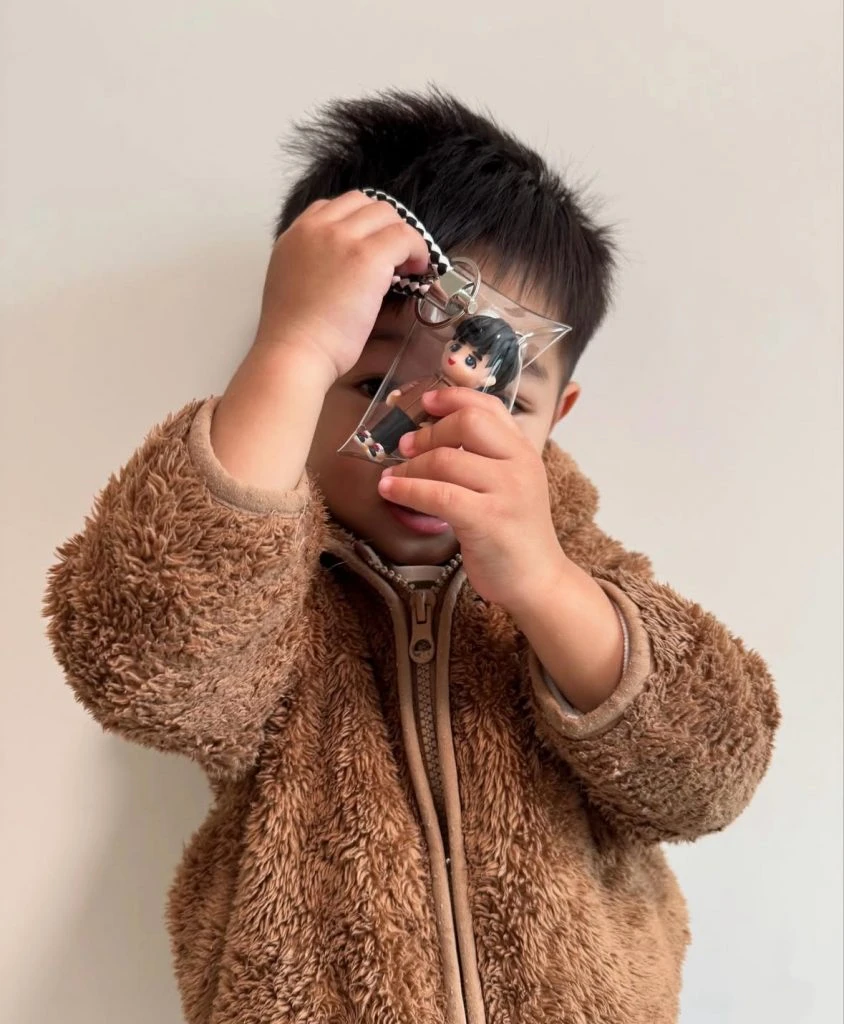 近距離視角，小朋友雙手高舉透明吊袋，袋中可見同款Q版人像公仔與金屬圈扣，毛絨外套質感清晰。