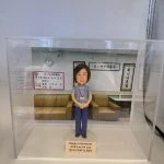 透明展示盒中的醫生人像公仔：角色穿藍灰色上衣與深藍長褲，掛識別證雙手於身前站立，置於淺色圓形底座。背板呈現門診等候區，有淺棕色沙發、牆上時鐘與中文標牌，左側公告板顯示日期與資訊。底座前方金色銘牌上寫有「Happy retirement APN Lee Po Lan QEH CGAT & GDH」。
