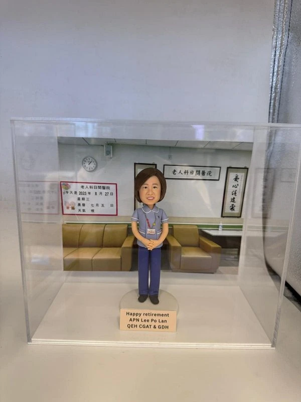 透明展示盒中的醫生人像公仔：角色穿藍灰色上衣與深藍長褲，掛識別證雙手於身前站立，置於淺色圓形底座。背板呈現門診等候區，有淺棕色沙發、牆上時鐘與中文標牌，左側公告板顯示日期與資訊。底座前方金色銘牌上寫有「Happy retirement APN Lee Po Lan QEH CGAT & GDH」。