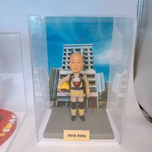 透明展示盒內擺放一個穿卡其黃色消防制服的人像公仔，胸前有呼吸器造型配件，手臂抱著黃色安全帽，腳穿黑色靴子。公仔站在灰色底座上，前方金色銘牌刻有「消防員」與姓名字樣。背景照片是藍天之下的多層白色建築與城市高樓，襯托消防主題氛圍。