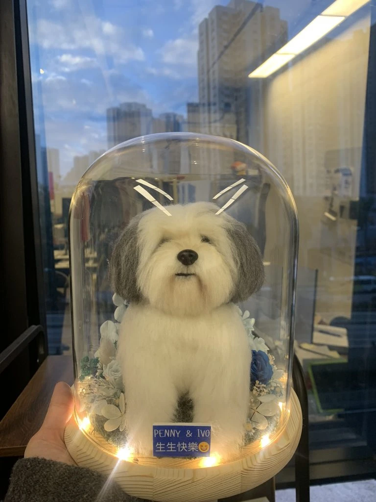 一個逼真嘅白色毛茸茸古代牧羊犬公仔，被玻璃罩和 LED 燈環繞，底部有「PENNY & IVO 生生快樂」字樣，展現精湛羊毛氈工藝，係訂造人像公仔嘅巔峰之作。