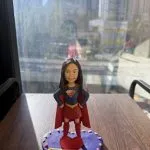 一尊以 女超人（Super Girl） 為主題的人像公仔作品。 中央公仔穿著紅色披風及深藍色戰衣，胸前綴有金紅色「S」標誌，擺出自信英勇姿勢。 面容細緻刻畫、笑容自然，五官比例與真人高度相似。 公仔立於一個紫色圓形底座上，邊緣裝飾以粉嫩心形點綴，前方放置紅色心形裝飾。 底座附有個人化訊息牌，上刻「You made chaos look classy. Now go rule another galaxy. All the best Super Jenny」。 鏡面背景映出窗外城市景觀，整體構圖充滿陽光光影與英氣氛圍。 作品象徵團隊中勇敢與靈感的精神，並作為送別同事的 Farewell紀念公仔。