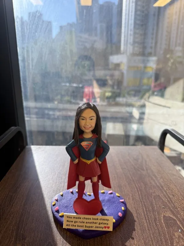 一尊以 女超人（Super Girl） 為主題的人像公仔作品。 中央公仔穿著紅色披風及深藍色戰衣，胸前綴有金紅色「S」標誌，擺出自信英勇姿勢。 面容細緻刻畫、笑容自然，五官比例與真人高度相似。 公仔立於一個紫色圓形底座上，邊緣裝飾以粉嫩心形點綴，前方放置紅色心形裝飾。 底座附有個人化訊息牌，上刻「You made chaos look classy. Now go rule another galaxy. All the best Super Jenny」。 鏡面背景映出窗外城市景觀，整體構圖充滿陽光光影與英氣氛圍。 作品象徵團隊中勇敢與靈感的精神，並作為送別同事的 Farewell紀念公仔。