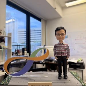 一隻手托著一座桌面模型：右側站立一個穿紅黑格紋襯衫、黑長褲與黑鞋的人像公仔，雙手插袋微笑；戴黑框眼鏡、短髮。左側為透明支架承托的八達通（Octopus）LOGO造型標誌，採橙、綠、紫三色帶狀設計。底座是灰色磚紋步道，兩側有綠色花草邊帶，前方金色銘牌寫著「YC Happy Retirement」。背景為室內辦公室與靠窗城市景色。