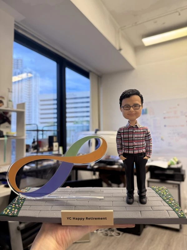 一隻手托著一座桌面模型：右側站立一個穿紅黑格紋襯衫、黑長褲與黑鞋的人像公仔，雙手插袋微笑；戴黑框眼鏡、短髮。左側為透明支架承托的八達通（Octopus）LOGO造型標誌，採橙、綠、紫三色帶狀設計。底座是灰色磚紋步道，兩側有綠色花草邊帶，前方金色銘牌寫著「YC Happy Retirement」。背景為室內辦公室與靠窗城市景色。