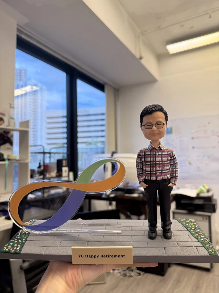 一隻手托著一座桌面模型：右側站立一個穿紅黑格紋襯衫、黑長褲與黑鞋的人像公仔，雙手插袋微笑；戴黑框眼鏡、短髮。左側為透明支架承托的八達通（Octopus）LOGO造型標誌，採橙、綠、紫三色帶狀設計。底座是灰色磚紋步道，兩側有綠色花草邊帶，前方金色銘牌寫著「YC Happy Retirement」。背景為室內辦公室與靠窗城市景色。