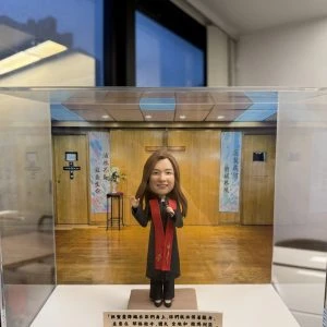 透明展示盒內的一尊人像公仔站在方形底座上，背景是木質牆面與十字架的教堂式佈景，左右各有彩色垂掛布幅與花台。公仔為長髮造型，穿深色禮服外搭紅色披帶，右手微舉、左手持麥克風，腳穿黑色鞋。展示盒前方有一條長形金色銘牌，上以黑字寫有多行中文祝福語。外部可見室內窗景與天花板燈光反射。