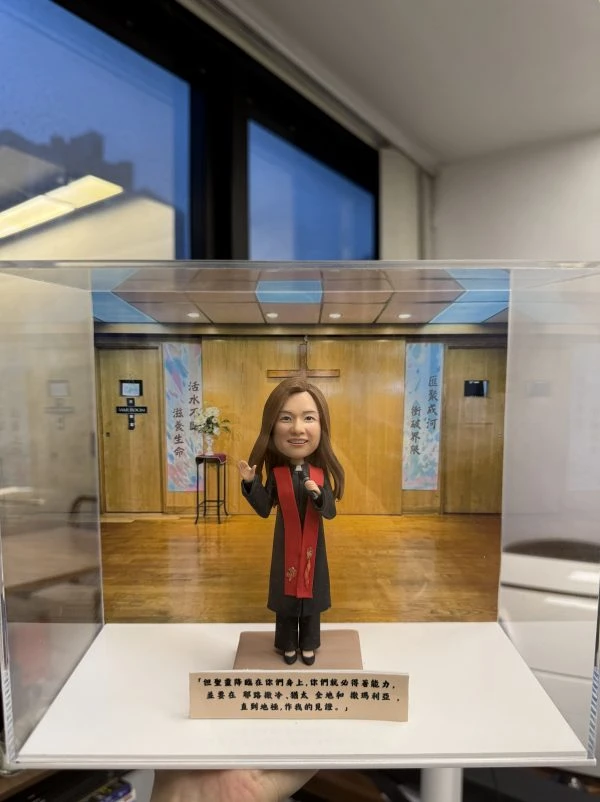 透明展示盒內的一尊人像公仔站在方形底座上，背景是木質牆面與十字架的教堂式佈景，左右各有彩色垂掛布幅與花台。公仔為長髮造型，穿深色禮服外搭紅色披帶，右手微舉、左手持麥克風，腳穿黑色鞋。展示盒前方有一條長形金色銘牌，上以黑字寫有多行中文祝福語。外部可見室內窗景與天花板燈光反射。