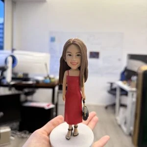 一隻精緻的人像公仔站在淺灰圓形底座上，被手掌托起展示；公仔為長直髮女性造型，穿紅色細肩帶連身裙與黑色厚底高跟鞋，右手拎著黑色小包。背景是開放式辦公室，可見桌面、螢幕、白板與文件，景深虛化讓主體更突出。