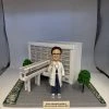 多張照片展示一組醫療主題的人像公仔場景：穿白袍、戴眼鏡的公仔站在灰色地磚平台上，頸掛證件、右手舉起示意。背後是白色方格磚紋的大型建築模型，立面有橫向百葉結構，前方連接一段高架走道與入口雨棚；平台兩側種有綠色圓頂小樹與鋪滿花瓣的綠化帶。前方小銘牌以中文寫上致謝字樣。部分照片顯示整套作品收納於透明展示盒，綁有綠色緞帶。