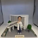 多張照片展示一組醫療主題的人像公仔場景：穿白袍、戴眼鏡的公仔站在灰色地磚平台上，頸掛證件、右手舉起示意。背後是白色方格磚紋的大型建築模型，立面有橫向百葉結構，前方連接一段高架走道與入口雨棚；平台兩側種有綠色圓頂小樹與鋪滿花瓣的綠化帶。前方小銘牌以中文寫上致謝字樣。部分照片顯示整套作品收納於透明展示盒，綁有綠色緞帶。