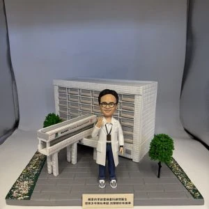 多張照片展示一組醫療主題的人像公仔場景：穿白袍、戴眼鏡的公仔站在灰色地磚平台上，頸掛證件、右手舉起示意。背後是白色方格磚紋的大型建築模型，立面有橫向百葉結構，前方連接一段高架走道與入口雨棚；平台兩側種有綠色圓頂小樹與鋪滿花瓣的綠化帶。前方小銘牌以中文寫上致謝字樣。部分照片顯示整套作品收納於透明展示盒，綁有綠色緞帶。