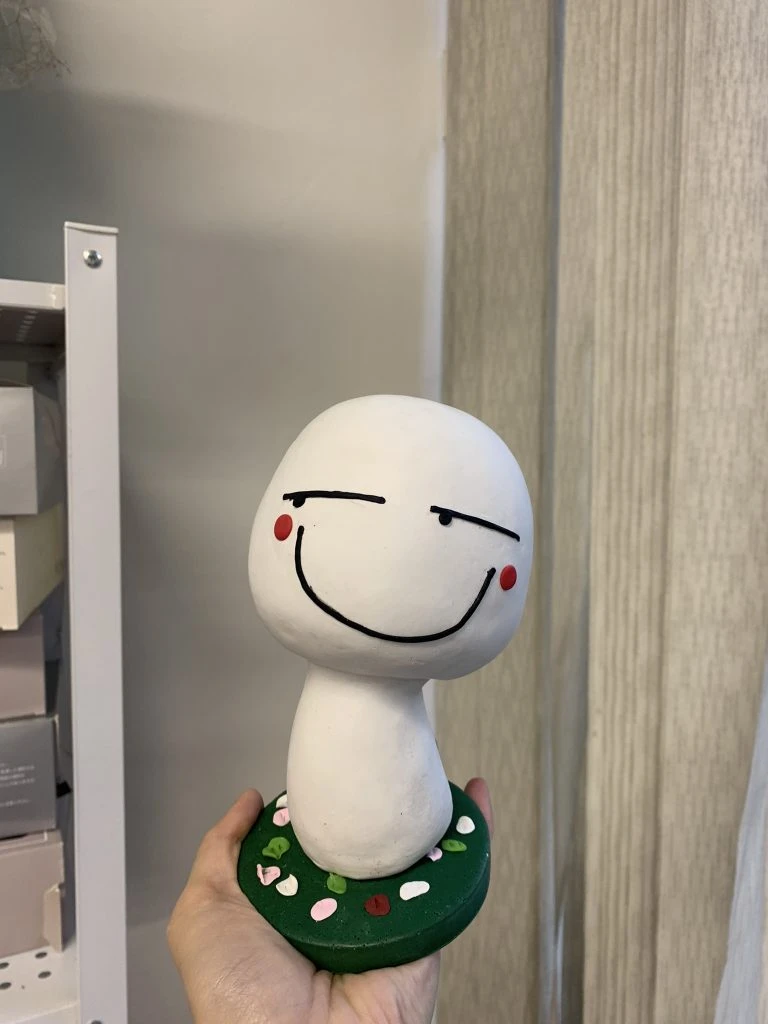 白色人形公仔，擁有卡通化的大頭和瞇眼笑容，臉頰兩邊有紅色腮紅，站在綠色圓形底座上，是訂造卡通人物公仔的趣味作品。