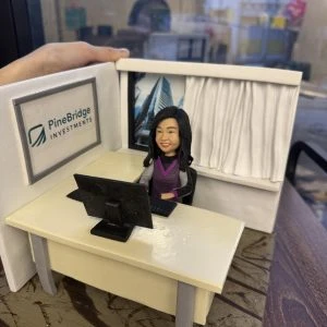 一個辦公室情境的人像公仔坐在米色辦公桌後方，面前有黑色螢幕與鍵盤；背牆左側掛有「PineBridge Investments」標誌框，右側為有白色窗簾與高樓外景的窗戶。公仔留中長髮、穿紫灰色上衣，帶微笑神情。整體擺在木紋底座上，左上角可見手掌扶著場景模型的邊框，背景為玻璃窗外的城市景色。