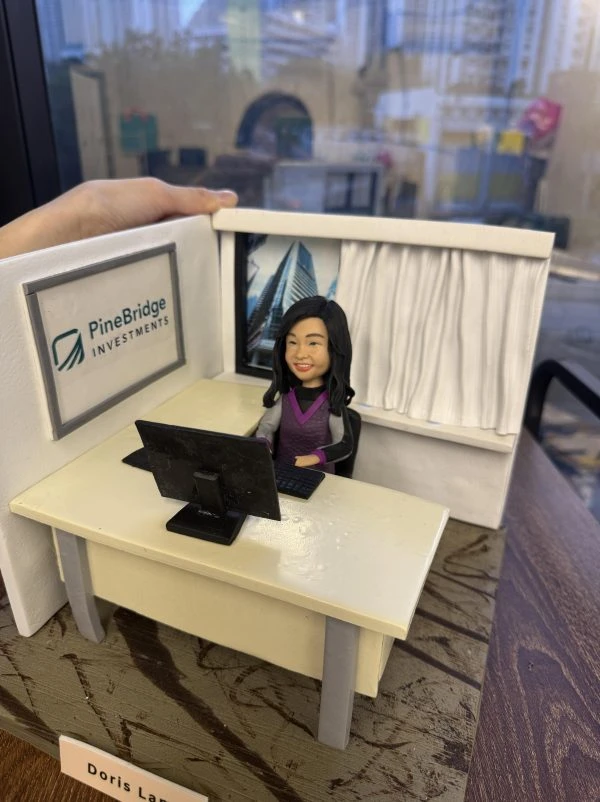 一個辦公室情境的人像公仔坐在米色辦公桌後方，面前有黑色螢幕與鍵盤；背牆左側掛有「PineBridge Investments」標誌框，右側為有白色窗簾與高樓外景的窗戶。公仔留中長髮、穿紫灰色上衣，帶微笑神情。整體擺在木紋底座上，左上角可見手掌扶著場景模型的邊框，背景為玻璃窗外的城市景色。