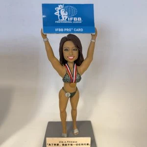 一款健身比基尼賽事風格的人像公仔站在灰色底座上，雙手高舉藍色牌匾，上印「IFBB PROFESSIONAL LEAGUE / IFBB PRO CARD」字樣。公仔著深綠比基尼與高跟舞台鞋，胸前掛著紅白藍項帶的金色獎牌，腰間有「1」號圓牌，表情自信微笑。底座前方有小銘牌，上有品牌與激勵語。背景為簡潔的白色牆面與桌面。