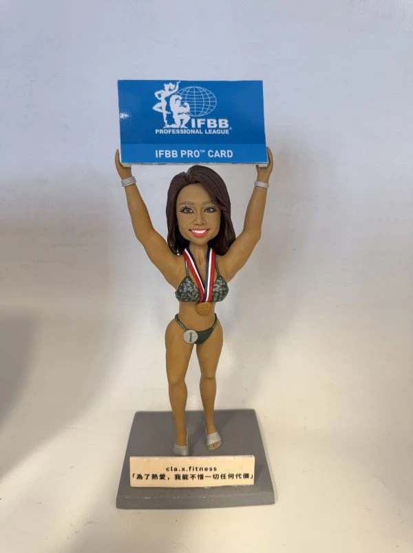 一款健身比基尼賽事風格的人像公仔站在灰色底座上，雙手高舉藍色牌匾，上印「IFBB PROFESSIONAL LEAGUE / IFBB PRO CARD」字樣。公仔著深綠比基尼與高跟舞台鞋，胸前掛著紅白藍項帶的金色獎牌，腰間有「1」號圓牌，表情自信微笑。底座前方有小銘牌，上有品牌與激勵語。背景為簡潔的白色牆面與桌面。