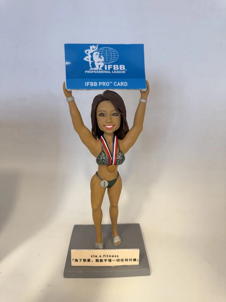 一款健身比基尼賽事風格的人像公仔站在灰色底座上，雙手高舉藍色牌匾，上印「IFBB PROFESSIONAL LEAGUE / IFBB PRO CARD」字樣。公仔著深綠比基尼與高跟舞台鞋，胸前掛著紅白藍項帶的金色獎牌，腰間有「1」號圓牌，表情自信微笑。底座前方有小銘牌，上有品牌與激勵語。背景為簡潔的白色牆面與桌面。
