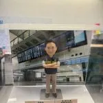 一款人像公仔置於透明展示盒內，背景為機場航班資訊顯示板與候機大廳結構。公仔穿深色背心式制服與卡其色長褲，胸前有徽章與「CUSTOMS」字樣臂章，雙臂交叉站立於灰色底座。展示盒前方放有名牌與獎牌式小牌座，顯示職稱與表揚訊息，整體呈現祝賀升遷的紀念氛圍。