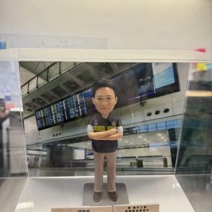 一款人像公仔置於透明展示盒內，背景為機場航班資訊顯示板與候機大廳結構。公仔穿深色背心式制服與卡其色長褲，胸前有徽章與「CUSTOMS」字樣臂章，雙臂交叉站立於灰色底座。展示盒前方放有名牌與獎牌式小牌座，顯示職稱與表揚訊息，整體呈現祝賀升遷的紀念氛圍。