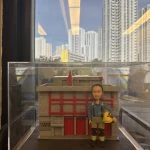 一座透明展示盒內的微縮場景：背景是灰色外牆、紅色直條門片與小型紅旗的建築模型，正面類似消防局外觀。前景站立一個穿藍灰上衣、卡其與黑色拼接褲、黑靴的人像公仔，肩有吊帶，右手提著黃色安全帽。底座前方有金色銘牌寫有祝賀退休字樣。展示盒置於室內窗邊，窗外可見高樓大廈與藍天。