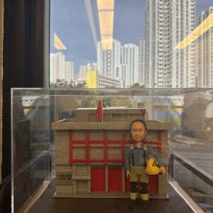 一座透明展示盒內的微縮場景:背景是灰色外牆、紅色直條門片與小型紅旗的建築模型,正面類似消防局外觀。前景站立一個穿藍灰上衣、卡其與黑色拼接褲、黑靴的人像公仔,肩有吊帶,右手提著黃色安全帽。底座前方有金色銘牌寫有祝賀退休字樣。展示盒置於室內窗邊,窗外可見高樓大廈與藍天。