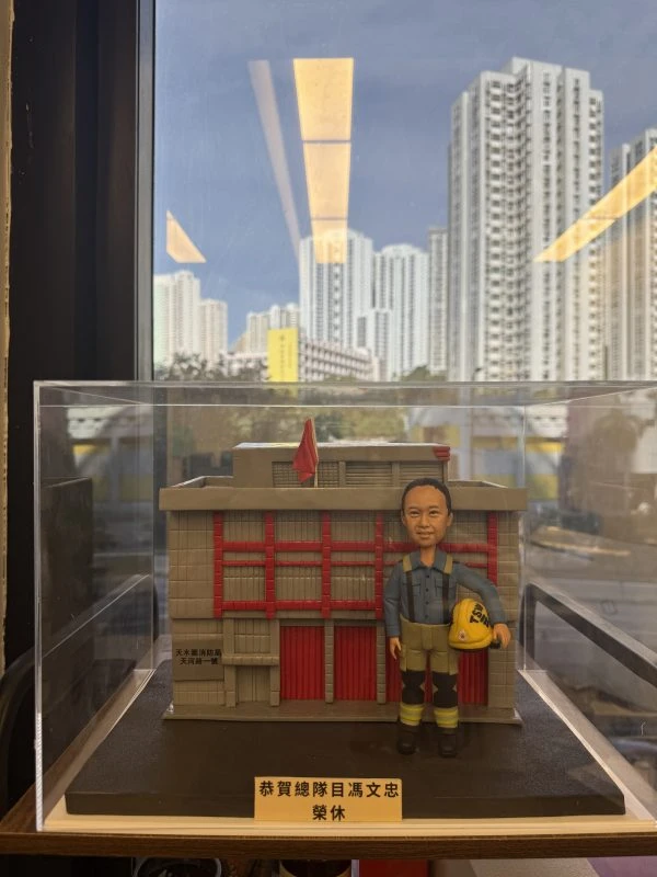 一座透明展示盒內的微縮場景：背景是灰色外牆、紅色直條門片與小型紅旗的建築模型，正面類似消防局外觀。前景站立一個穿藍灰上衣、卡其與黑色拼接褲、黑靴的人像公仔，肩有吊帶，右手提著黃色安全帽。底座前方有金色銘牌寫有祝賀退休字樣。展示盒置於室內窗邊，窗外可見高樓大廈與藍天。
