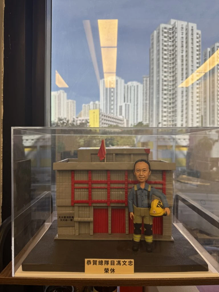 一座透明展示盒內的微縮場景：背景是灰色外牆、紅色直條門片與小型紅旗的建築模型，正面類似消防局外觀。前景站立一個穿藍灰上衣、卡其與黑色拼接褲、黑靴的人像公仔，肩有吊帶，右手提著黃色安全帽。底座前方有金色銘牌寫有祝賀退休字樣。展示盒置於室內窗邊，窗外可見高樓大廈與藍天。