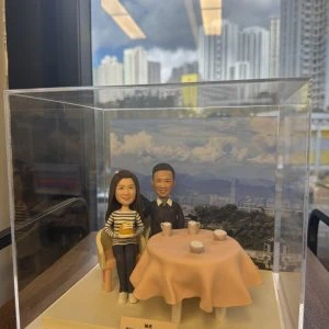 透明壓克力展示盒中的雙人退休人像公仔：左側角色穿條紋上衣、黑褲與白鞋，雙手捧杯入席；右側角色著深色西裝，並肩坐在圓桌旁。桌面覆粉橘色桌布，擺放三只茶杯與一只小盅，呈現酒樓用茶氣氛。前方小銘牌寫有「Happy Retirement 2025」與「Happy birthday 2025」。背板為城市高樓與遠山景致，整體置於木質檯面。