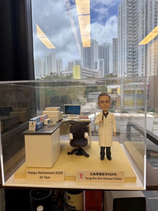 一個放在透明壓克力盒內的牙科主題場景：穿白色醫師袍的牙醫人像公仔站在米黃色底座上，旁邊是深色診療椅與白色診療檯，檯面擺有文件盒、口罩盒與工具罐。右前角的金色銘牌寫著「Happy Retirement 2025 Dr Tam」，側邊垂直銘牌標示「Tang Siu Kin Dental Clinic 北區鄧兆堅牙科診所」。背景透過窗景可見高樓與藍天。