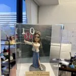 透明亞克力展示盒內的一款人像公仔：長髮、戴墨鏡的角色手持銀色「1」「0」氣球造型道具，身穿白色外套與開衩藍色長裙、運動鞋，站在灰色圓形底座上。前方金色銘牌刻有「Dear Cheryl Lululemon 10th anniversary」文字