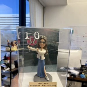 透明亞克力展示盒內的一款人像公仔:長髮、戴墨鏡的角色手持銀色「1」「0」氣球造型道具,身穿白色外套與開衩藍色長裙、運動鞋,站在灰色圓形底座上。前方金色銘牌刻有「Dear Cheryl Lululemon 10th anniversary」文字