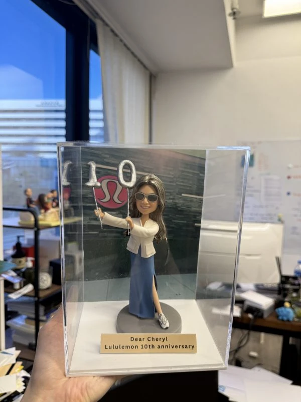 透明亞克力展示盒內的一款人像公仔：長髮、戴墨鏡的角色手持銀色「1」「0」氣球造型道具，身穿白色外套與開衩藍色長裙、運動鞋，站在灰色圓形底座上。前方金色銘牌刻有「Dear Cheryl Lululemon 10th anniversary」文字