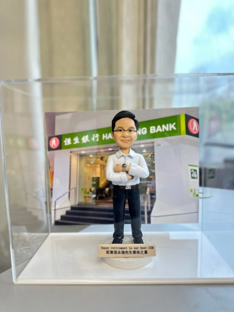 透明展示盒與桌面擺拍的兩款客製作品。背景均為恆生銀行門面照片：一張是玻璃帷幕與白色外牆、正中有紅圓徽與「恆生銀行 HANG SENG BANK」字樣；另一張是綠底白字的分行門頭。前景為綠色磚紋底座，其一作品擺放大型紅色球體立體LOGO（白色筆畫圖案），旁邊站著戴眼鏡、白襯衫黑長褲與棕色皮鞋的男性人像公仔，雙手叉腰；底座前有金屬色名牌寫上「David Wish you all the best! Jul 2024」。另一作品則為透明盒中展示，背景是分行入口，前方立著雙臂交叉、白襯衫深色長褲與手錶的男性人像公仔，底部名牌刻有退休祝福。兩款皆以恆生銀行門面為立體建築背景，強調品牌元素與個人化訊息。