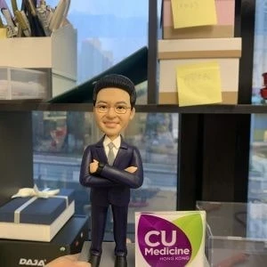 桌面擺設近景：一位穿深紫色西裝、白襯衫與領帶的人像公仔雙臂交叉站立，面帶微笑；右側是一個方形底座的立體立牌，前方為紫色葉形、後方為萊姆綠葉形，白字標示「CU Medicine HONG KONG」。背景是辦公室書架與窗外城市街景。