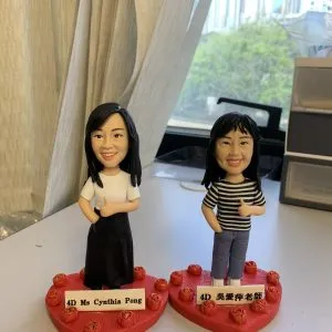 (客製化Q版人物公仔——雙人教師紀念擺飾 手工人物公仔細節（圖片展示了兩尊客製化的Q版人物黏土公仔，主角為兩位女性教師，並排站立。左側公仔留著黑色長直髮，身穿白色圓領T恤搭配黑色長裙；右側公仔留著齊肩短髮，穿著黑白條紋上衣與藍色長褲。兩者皆擺出右手插口袋、左手比讚的自信姿勢，面部表情刻畫細膩，展現出親切燦爛的笑容。公仔底部皆固定於紅色愛心形狀的底座上，底座邊緣裝飾著一圈帶有金粉點綴的紅色立體玫瑰花，前方分別立有白色名牌，黑字寫著「4D Ms Cynthia Pong」與「4D 吳愛萍老師」） 環境背景與光影（公仔放置於室內靠窗的桌面上進行拍攝，左側有米白色的窗簾垂下，右側可見部分辦公收納櫃。透過後方的玻璃窗，可以看見戶外的城市高樓建築與綠樹，顯示出自然光線充足的環境。自然光從後方與側面灑入，使公仔臉部與身體呈現立體的光影變化，背景的生活化場景增添了真實感與溫馨的氛圍） 軟陶公仔、真人公仔訂製、Q版人像、謝師宴禮物、教師節禮物、手工捏塑、客製化名牌、職業公仔、愛心底座、輕黏土手作 畢業紀念、感謝師恩、同事升遷禮物、辦公桌擺飾、獨家訂製紀念品、退休禮物、活動贈禮)