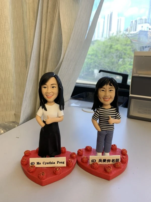 (客製化Q版人物公仔——雙人教師紀念擺飾 手工人物公仔細節（圖片展示了兩尊客製化的Q版人物黏土公仔，主角為兩位女性教師，並排站立。左側公仔留著黑色長直髮，身穿白色圓領T恤搭配黑色長裙；右側公仔留著齊肩短髮，穿著黑白條紋上衣與藍色長褲。兩者皆擺出右手插口袋、左手比讚的自信姿勢，面部表情刻畫細膩，展現出親切燦爛的笑容。公仔底部皆固定於紅色愛心形狀的底座上，底座邊緣裝飾著一圈帶有金粉點綴的紅色立體玫瑰花，前方分別立有白色名牌，黑字寫著「4D Ms Cynthia Pong」與「4D 吳愛萍老師」） 環境背景與光影（公仔放置於室內靠窗的桌面上進行拍攝，左側有米白色的窗簾垂下，右側可見部分辦公收納櫃。透過後方的玻璃窗，可以看見戶外的城市高樓建築與綠樹，顯示出自然光線充足的環境。自然光從後方與側面灑入，使公仔臉部與身體呈現立體的光影變化，背景的生活化場景增添了真實感與溫馨的氛圍） 軟陶公仔、真人公仔訂製、Q版人像、謝師宴禮物、教師節禮物、手工捏塑、客製化名牌、職業公仔、愛心底座、輕黏土手作 畢業紀念、感謝師恩、同事升遷禮物、辦公桌擺飾、獨家訂製紀念品、退休禮物、活動贈禮)
