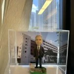 一個放在透明展示盒內的桌面人像公仔。公仔為短灰髮、微笑表情的男性形象，穿深色西裝與白襯衫、紅黑條紋領帶，右手自然下垂、左手插袋，站姿端正。腳下是綠色草地造型底座，左前方有籐籃與綠植裝飾，前側白色牌子印有中文感謝與祝福字樣。背板是一棟多層校舍的照片，可見立面、窗戶與棕櫚樹，牆上有中文校名。展示盒由手掌托起，背景是室內窗邊、米色窗簾與窗外高樓、藍天白雲。