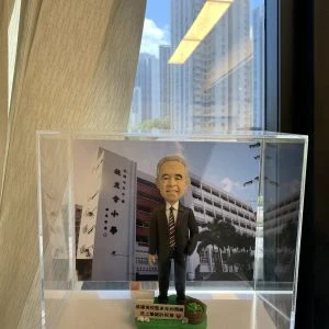 一個放在透明展示盒內的桌面人像公仔。公仔為短灰髮、微笑表情的男性形象,穿深色西裝與白襯衫、紅黑條紋領帶,右手自然下垂、左手插袋,站姿端正。腳下是綠色草地造型底座,左前方有籐籃與綠植裝飾,前側白色牌子印有中文感謝與祝福字樣。背板是一棟多層校舍的照片,可見立面、窗戶與棕櫚樹,牆上有中文校名。展示盒由手掌托起,背景是室內窗邊、米色窗簾與窗外高樓、藍天白雲。