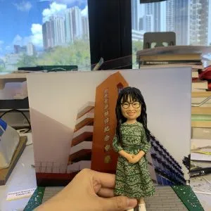 (客製化Q版女教師公仔——校園背景榮休紀念擺飾 手工人物公仔細節(圖片展示了一尊客製化的Q版女性人物黏土公仔,主角留著黑色長捲髮並有瀏海,戴著黑框眼鏡,面帶親切微笑。她身穿一件綠色底白色花紋的長袖連身裙,腳踩白色休閒鞋,雙手優雅地交疊在身前。公仔最顯著的特點是其背景板,是一張「樂善堂梁植偉紀念中學」橙色校舍大樓的實景照片,還原了校園場景。公仔立於仿灰色磚石紋路的底座上,前方名牌寫著「春風化雨四十載 育人靈傑桃李滿園」,點明了這是一份紀念執教四十週年的榮休或長期服務禮物) 環境背景與光影(公仔被放置在一個看似工作檯的環境中,由一隻手扶持展示。背景稍顯雜亂,可見堆疊的書籍、文件及製作工具,暗示這是在製作完成後的檢視環節。後方是大片落地窗,窗外是藍天白雲與高樓大廈的城市景觀,明亮的自然光線照射進來,使公仔的色彩鮮豔,綠色裙裝與橙色校舍背景形成鮮明對比) 軟陶公仔、真人公仔訂製、Q版人像、女教師禮物、校長禮物、榮休紀念、校園場景還原、綠色碎花裙、樂善堂梁植偉紀念中學、長期服務獎 感謝師恩、四十週年紀念、教育工作者、獨家訂製、辦公室擺設、移居紀念、桃李滿門、手工藝品)