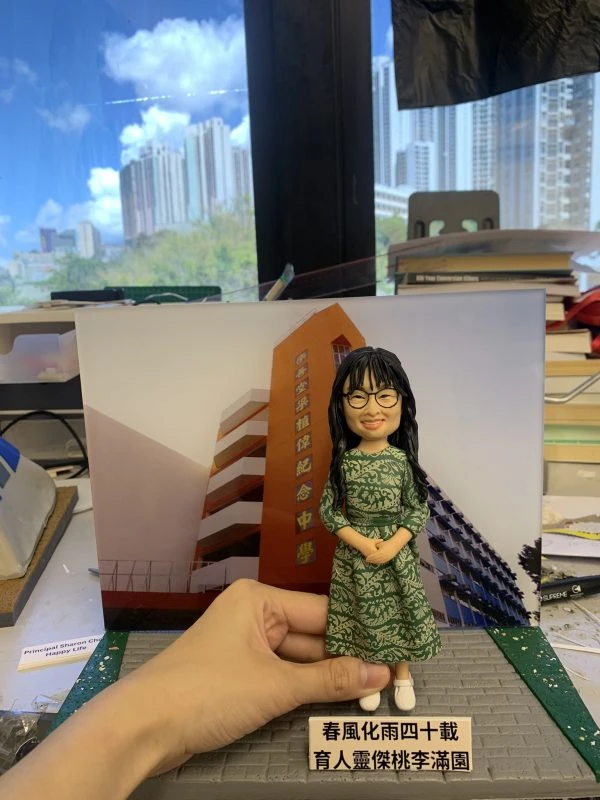 (客製化Q版女教師公仔——校園背景榮休紀念擺飾 手工人物公仔細節（圖片展示了一尊客製化的Q版女性人物黏土公仔，主角留著黑色長捲髮並有瀏海，戴著黑框眼鏡，面帶親切微笑。她身穿一件綠色底白色花紋的長袖連身裙，腳踩白色休閒鞋，雙手優雅地交疊在身前。公仔最顯著的特點是其背景板，是一張「樂善堂梁植偉紀念中學」橙色校舍大樓的實景照片，還原了校園場景。公仔立於仿灰色磚石紋路的底座上，前方名牌寫著「春風化雨四十載 育人靈傑桃李滿園」，點明了這是一份紀念執教四十週年的榮休或長期服務禮物） 環境背景與光影（公仔被放置在一個看似工作檯的環境中，由一隻手扶持展示。背景稍顯雜亂，可見堆疊的書籍、文件及製作工具，暗示這是在製作完成後的檢視環節。後方是大片落地窗，窗外是藍天白雲與高樓大廈的城市景觀，明亮的自然光線照射進來，使公仔的色彩鮮豔，綠色裙裝與橙色校舍背景形成鮮明對比） 軟陶公仔、真人公仔訂製、Q版人像、女教師禮物、校長禮物、榮休紀念、校園場景還原、綠色碎花裙、樂善堂梁植偉紀念中學、長期服務獎 感謝師恩、四十週年紀念、教育工作者、獨家訂製、辦公室擺設、移居紀念、桃李滿門、手工藝品)